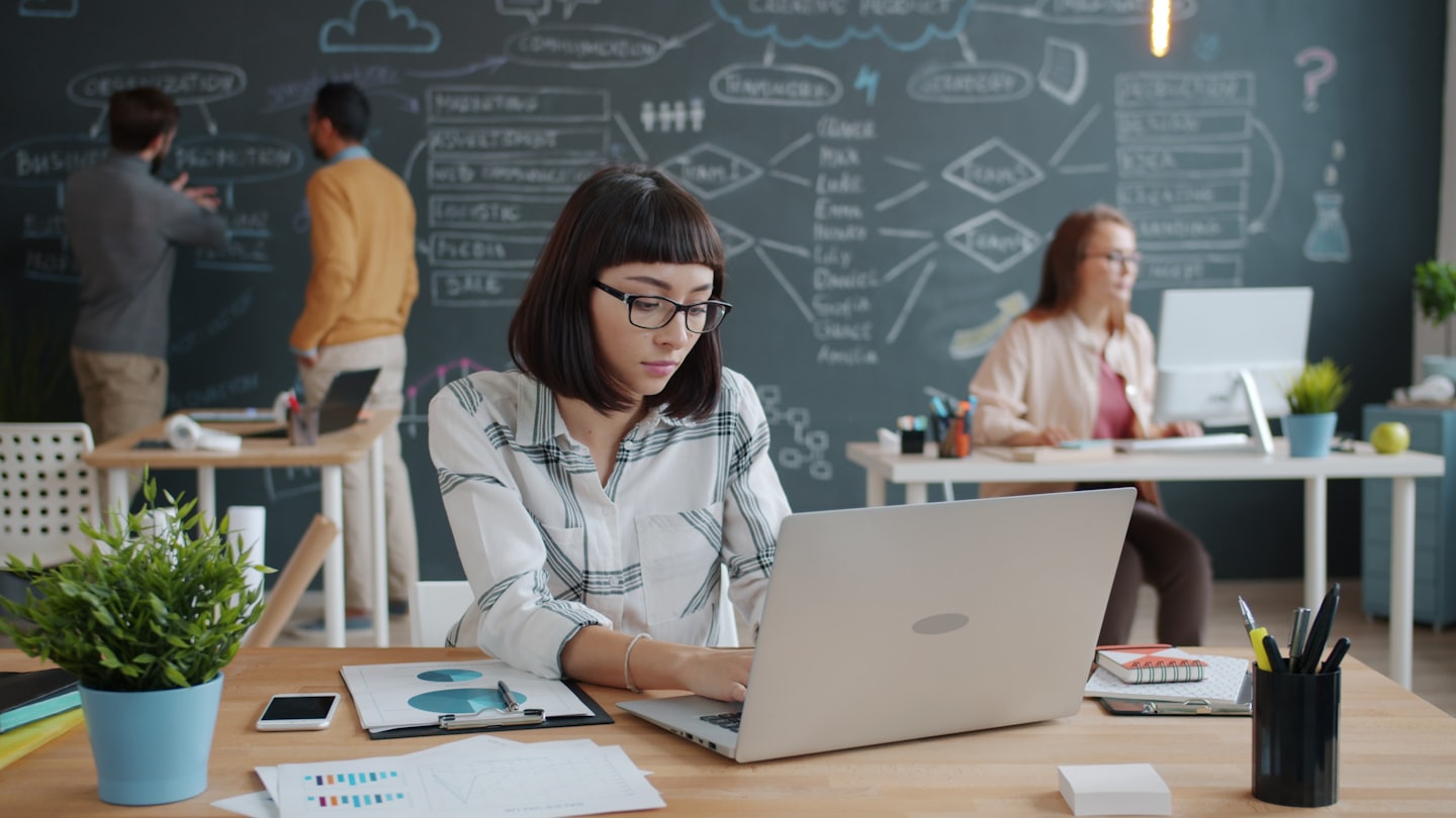 Woman wearing glasses works intently on laptop at wooden desk.