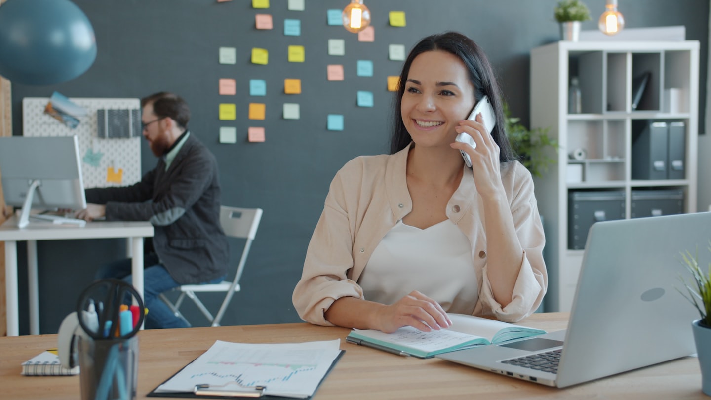 Smiling woman talks on smartphone at desk with open notebook.
